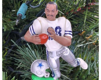 May include: A Christmas tree ornament depicting a football player in a white and blue uniform, holding a red football. A silver helmet with a blue star rests on a green base. The ornament is hanging on a green Christmas tree.