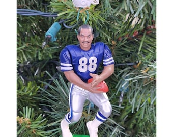 May include: A Christmas tree ornament featuring a football player in a blue and white uniform with the number 88. The player is holding a red football. A white helmet with a blue star is above the player.