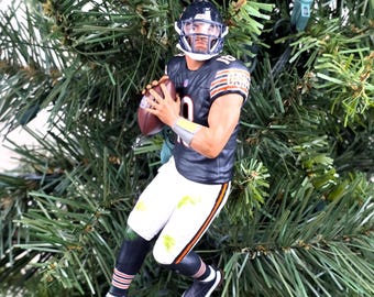 Może przedstawiać: Figurka zawodnika futbolu amerykańskiego Chicago Bears jako ozdoba, ubrana w granatową koszulkę z numerem 10, białe spodnie w pomarańczowo-granatowe paski, granatowy kask z maską, trzymająca brązową piłkę, zawieszona na zielonym sznurku.