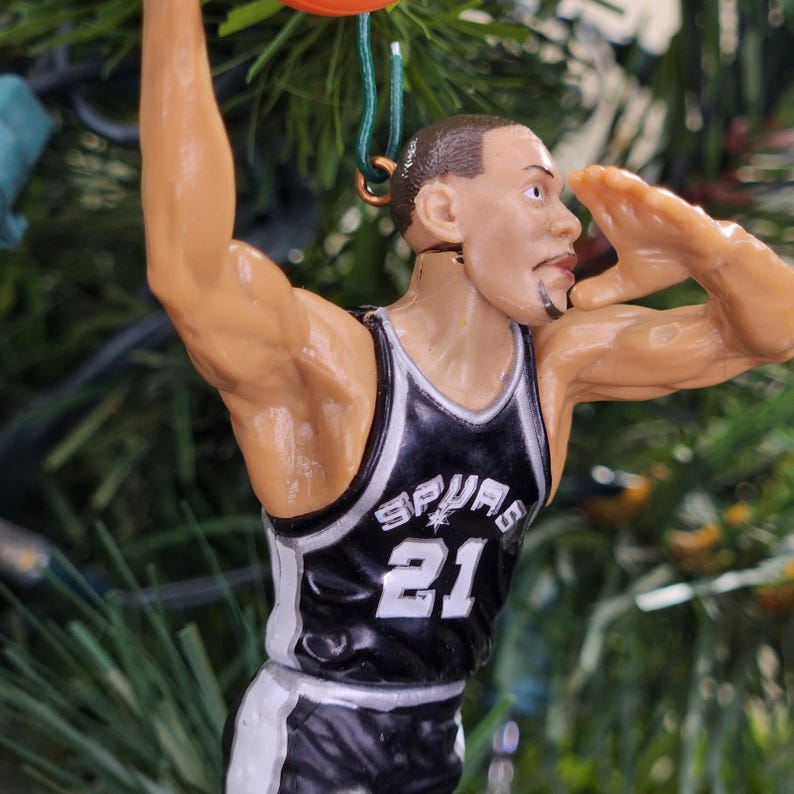 May include: A figurine of a basketball player in a black and white jersey with the number 21. The figurine is holding a basketball and has a detailed facial expression. The word "SPURS" is visible on the jersey.