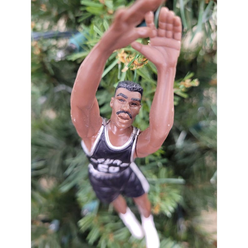 May include: A figurine of a basketball player in a black and white uniform, arms raised. The figure is wearing a jersey with the text "SPURS" and black shorts. The figure is a Christmas tree ornament.