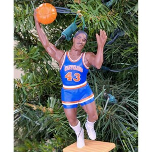 May include: A basketball-themed Christmas ornament featuring a figurine of a basketball player in a blue and white Cleveland jersey with the number 43. The player is holding an orange basketball. The ornament is attached to a green Christmas tree.