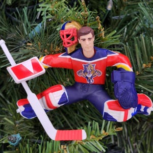 May include: A hockey goalie Christmas ornament. The ornament is in a goalie stance, wearing a red, blue, and white uniform with a team logo. The goalie has a mask and a hockey stick. The ornament is on a green Christmas tree.