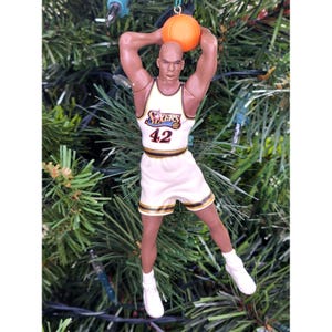 May include: A basketball-themed Christmas ornament featuring a bald player in a white and gold Sixers jersey with the number 42. The player is holding an orange basketball above their head. The ornament is set against a backdrop of green Christmas tree branches.
