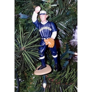 May include: A baseball-themed Christmas tree ornament. The figurine depicts a baseball player in a navy blue uniform with the word "Washington" on the chest, a tan glove, and a white baseball. The ornament is attached to a green Christmas tree.