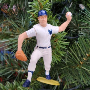 May include: A baseball player figurine in a white uniform with blue accents, a blue cap, and a brown glove. The figurine is holding a baseball and is positioned as if throwing a pitch. The figurine is on a yellow base.
