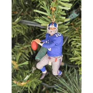 May include: A miniature football player ornament in a blue jersey with the number 10, gray pants, and a white helmet. The player is holding a red football. The ornament is hanging on a Christmas tree.