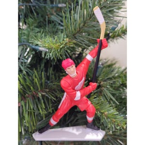 May include: A red hockey player figurine in a dynamic pose, holding a hockey stick with a white blade. The player wears a red uniform with white accents and a red cap. The figurine is mounted on a white base, perfect for holiday decor.