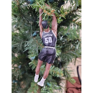 May include: A Christmas tree ornament depicting a basketball player in a black and white jersey with the name "Robinson" and the number "50". The ornament is hanging from a green Christmas tree.