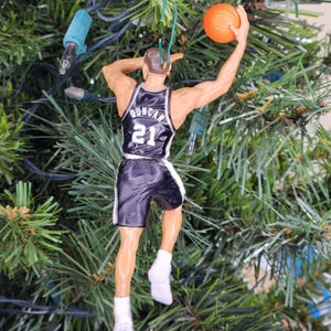 May include: A basketball-themed Christmas tree ornament. The ornament depicts a basketball player in a black and white uniform with the name "Duncan" and the number "21" on the jersey. The player is in mid-air, holding an orange basketball.