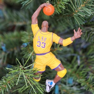 May include: A yellow and purple LSU basketball player ornament in mid-air, holding an orange basketball. The figure is wearing a yellow jersey and shorts with the number 33. The ornament is hanging from a gold hook on a Christmas tree.