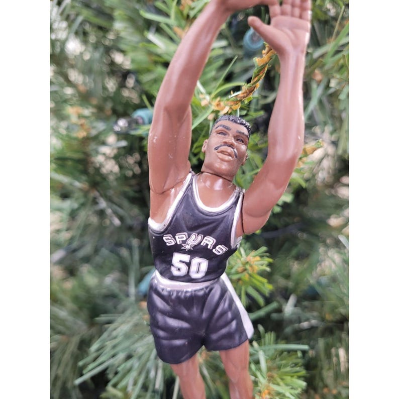 May include: A figurine of a basketball player in a black and white Spurs uniform, number 50, with arms raised. The figure is a Christmas tree ornament. The background is a blurred green.