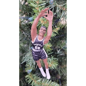 May include: A basketball player ornament in a black and white uniform with the number 50. The ornament is suspended in front of a green Christmas tree. The player is in a shooting pose with arms extended.