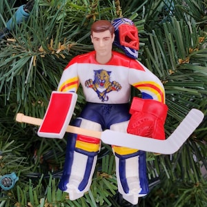 May include: A hockey goalie Christmas ornament. The ornament features a figure in a white, red, and blue jersey with a tiger logo, blue shorts, and goalie pads. A red helmet rests on the figure's head, and a hockey stick and glove are included.