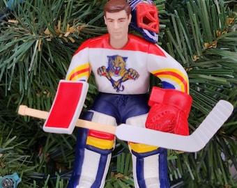 May include: A hockey goalie Christmas ornament. The ornament features a figure in a white, red, and blue jersey with a tiger logo, blue shorts, and goalie pads. A red helmet rests on the figure's head, and a hockey stick and glove are included.