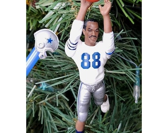May include: A football-themed Christmas tree ornament. The ornament depicts a football player in a white jersey with the number 88 in blue. The player is reaching for an orange football. A blue and white helmet is also visible.