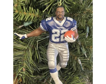 May include: A football-themed Christmas tree ornament. The ornament depicts a football player in a blue and silver uniform, holding an orange football. The player's jersey has the number 22. The ornament is hanging on a green Christmas tree.