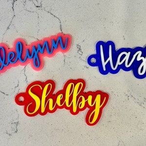 May include: Three personalized name tags, each with a different name. The name "Adelynn" is in blue lettering on a pink background. The name "Hazel" is in white lettering on a blue background. The name "Shelby" is in yellow lettering on a red background.