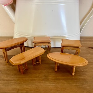 May include: A collection of miniature wooden tables in various shapes: oval, rectangular, and square. The tables are light brown with visible wood grain and are set against a neutral background. The tables are designed for a dollhouse or miniature setting.
