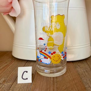 Op de afbeelding: Transparant glas met een gele Funshine Bear-afbeelding en de woorden "Funshine Bear" en "Feeling Fantastic". Het glas heeft ook een regenboog, een lachende zon en sterren. Een witte kan met roze bloemen staat op de achtergrond.