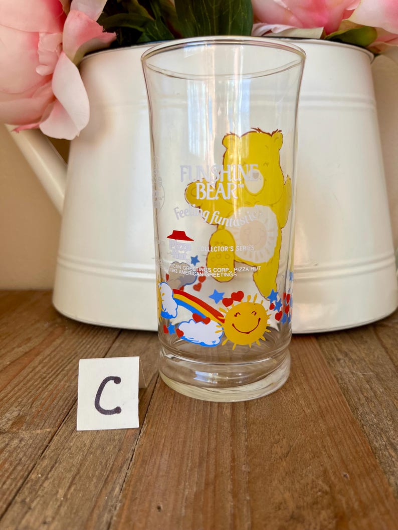 Op de afbeelding: Transparant glas met een gele Funshine Bear-afbeelding en de woorden "Funshine Bear" en "Feeling Fantastic". Het glas heeft ook een regenboog, een lachende zon en sterren. Een witte kan met roze bloemen staat op de achtergrond.
