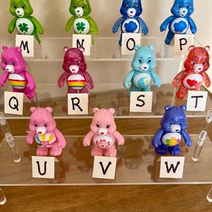 Puede incluir: Una colecci&oacute;n de doce figuras de osos amorosos en varios colores, incluyendo verde, rosa, azul y rojo. Cada oso tiene una insignia &uacute;nica en el vientre y se exhibe en una estanter&iacute;a de acr&iacute;lico transparente con etiquetas con letras.