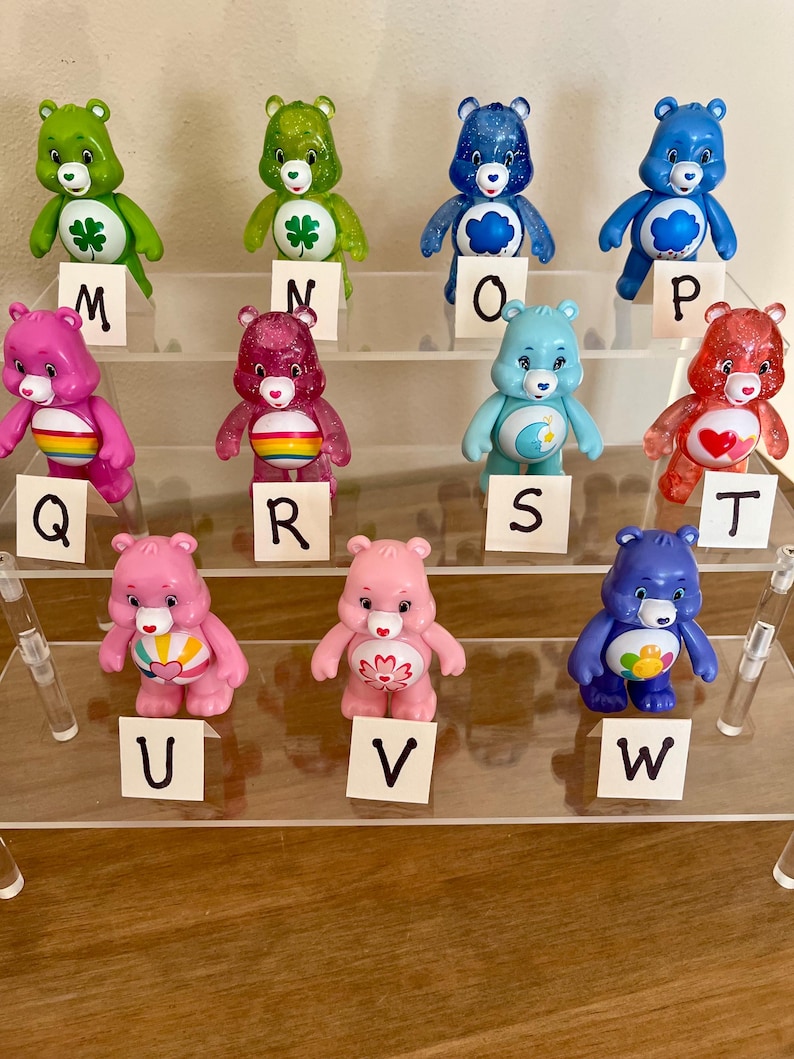 Puede incluir: Una colecci&oacute;n de doce figuras de osos amorosos en varios colores, incluyendo verde, rosa, azul y rojo. Cada oso tiene una insignia &uacute;nica en el vientre y se exhibe en una estanter&iacute;a de acr&iacute;lico transparente con etiquetas con letras.