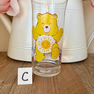 Op de afbeelding: Een helder glas met een gele Care Bear-afbeelding, met een lachende zon op de buik. De beer heeft zijn armen uitgestrekt. Het glas staat op een houten oppervlak met een witte kan en roze bloemen op de achtergrond. Een klein wit kaartje met de letter "C" ervoor.