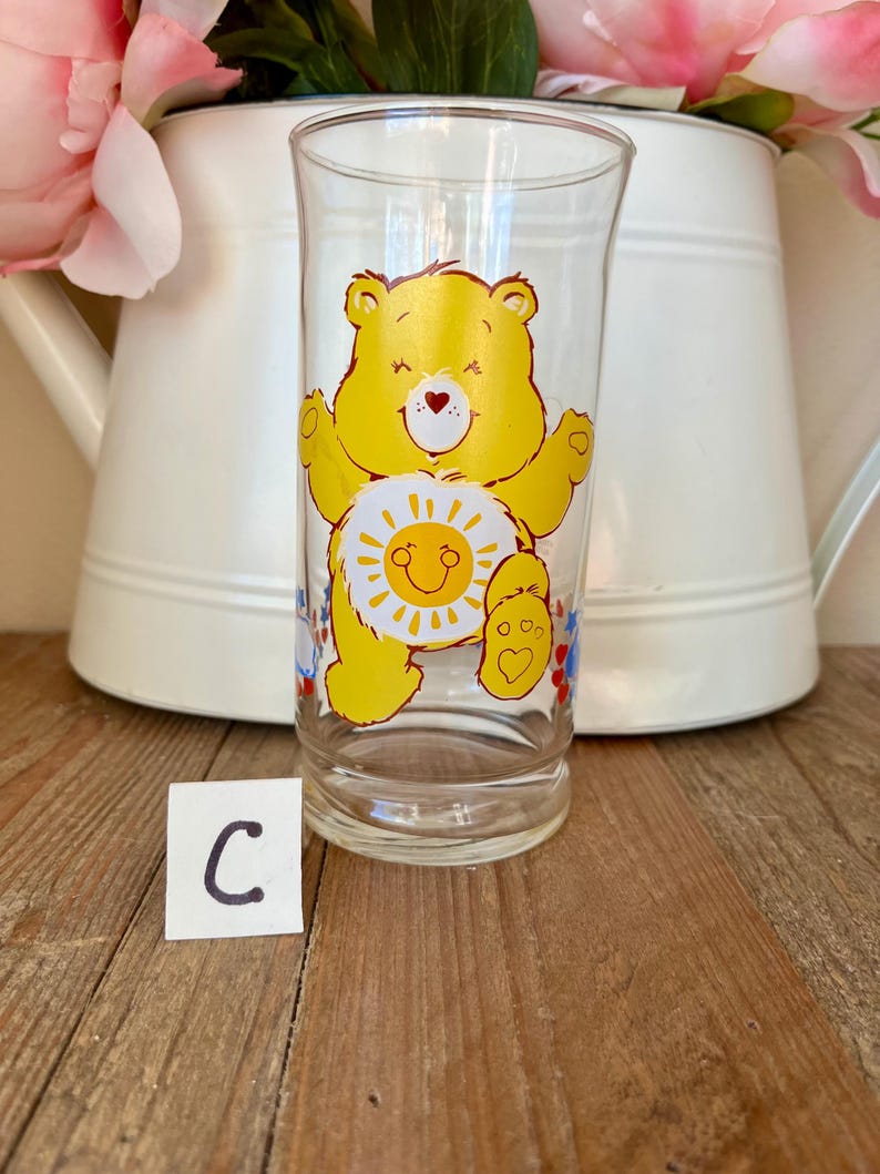 Op de afbeelding: Een helder glas met een gele Care Bear-afbeelding, met een lachende zon op de buik. De beer heeft zijn armen uitgestrekt. Het glas staat op een houten oppervlak met een witte kan en roze bloemen op de achtergrond. Een klein wit kaartje met de letter "C" ervoor.