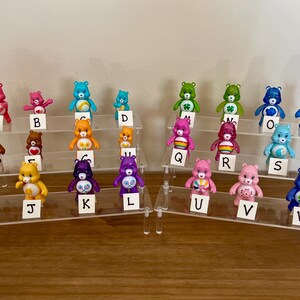 Puede incluir: Una colecci&oacute;n de coloridas figuras de Ositos Cari&ntilde;ositos, cada una con dise&ntilde;os y colores &uacute;nicos, dispuestas en expositores escalonados. Cada oso est&aacute; etiquetado con una letra del alfabeto, creando una exhibici&oacute;n l&uacute;dica y coleccionable. Los osos son de pl&aacute;stico.