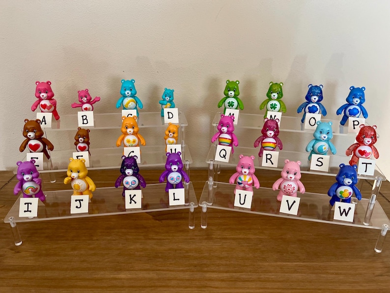 Puede incluir: Una colecci&oacute;n de coloridas figuras de Ositos Cari&ntilde;ositos, cada una con dise&ntilde;os y colores &uacute;nicos, dispuestas en expositores escalonados. Cada oso est&aacute; etiquetado con una letra del alfabeto, creando una exhibici&oacute;n l&uacute;dica y coleccionable. Los osos son de pl&aacute;stico.