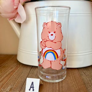 Op de afbeelding: Een helder glazen tumbler met een roze Care Bear-personage met een regenboog op zijn buik. Het glas heeft een rechte, cilindrische vorm en is versierd met kleurrijke Care Bear-afbeeldingen. De achtergrond omvat een witte kan en roze bloemen.