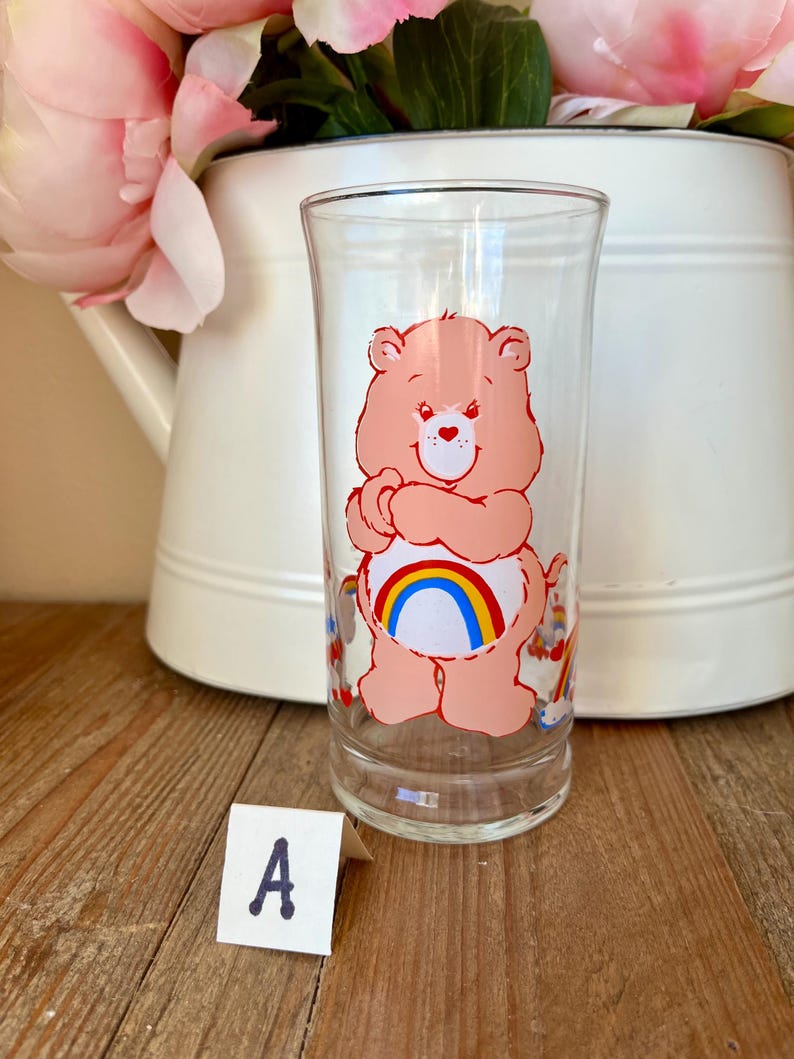 Op de afbeelding: Een helder glazen tumbler met een roze Care Bear-personage met een regenboog op zijn buik. Het glas heeft een rechte, cilindrische vorm en is versierd met kleurrijke Care Bear-afbeeldingen. De achtergrond omvat een witte kan en roze bloemen.
