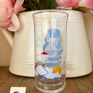 Op de afbeelding: Een helder glas met een blauw Grumpy Bear-ontwerp en de tekst "Hugs welcome". Het glas heeft een wolken- en zonontwerp aan de onderkant. De woorden "Collector's Series" zijn ook zichtbaar.