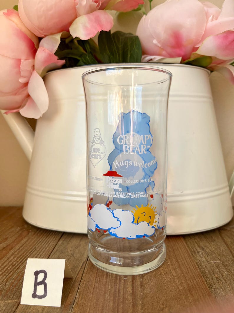 Op de afbeelding: Een helder glas met een blauw Grumpy Bear-ontwerp en de tekst "Hugs welcome". Het glas heeft een wolken- en zonontwerp aan de onderkant. De woorden "Collector's Series" zijn ook zichtbaar.