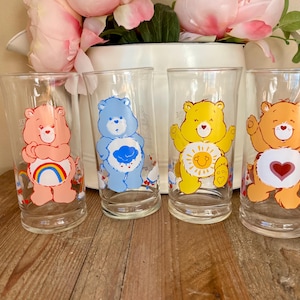 Op de afbeelding: Set van vier vintage Care Bear drinkglazen. Elk glas toont een ander Care Bear-personage in levendige kleuren: roze met een regenboog, blauw met een wolk, geel met een zon en oranje met een hart. De glazen zijn helder en cilindrisch.