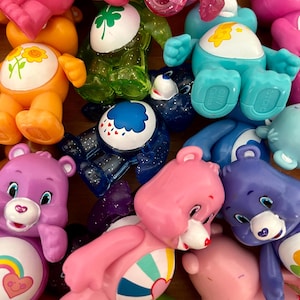 Puede incluir: Colecci&oacute;n de figuras de osos cari&ntilde;osos de colores en varias poses. Los osos son de color rosa, azul, naranja, verde y morado. Cada oso tiene una insignia &uacute;nica en el vientre, como un arco&iacute;ris, un coraz&oacute;n o una flor. Los juguetes son de pl&aacute;stico.