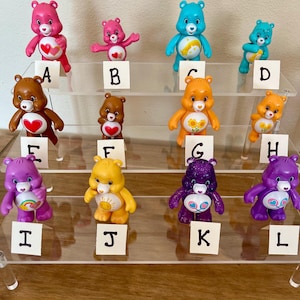 Puede incluir: Una colecci&oacute;n de doce figuras de ositos cari&ntilde;osos en varios colores, incluyendo rosa, azul, marr&oacute;n, naranja, amarillo y morado. Cada oso tiene una insignia &uacute;nica en el vientre y se exhibe en una estanter&iacute;a de acr&iacute;lico transparente. Los osos est&aacute;n etiquetados de la A a la L.