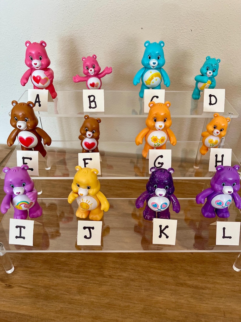 Puede incluir: Una colecci&oacute;n de doce figuras de ositos cari&ntilde;osos en varios colores, incluyendo rosa, azul, marr&oacute;n, naranja, amarillo y morado. Cada oso tiene una insignia &uacute;nica en el vientre y se exhibe en una estanter&iacute;a de acr&iacute;lico transparente. Los osos est&aacute;n etiquetados de la A a la L.