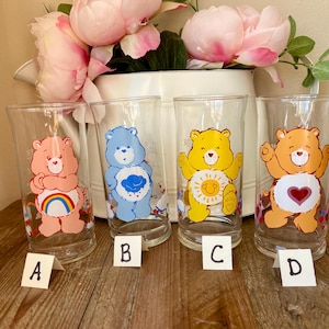 Op de afbeelding: Vier heldere glazen bekers met Care Bear-ontwerpen. Elk glas heeft een andere beer: roze met een regenboog, blauw met een wolk, geel met een zon en oranje met een hart. De glazen zijn gelabeld A, B, C en D.