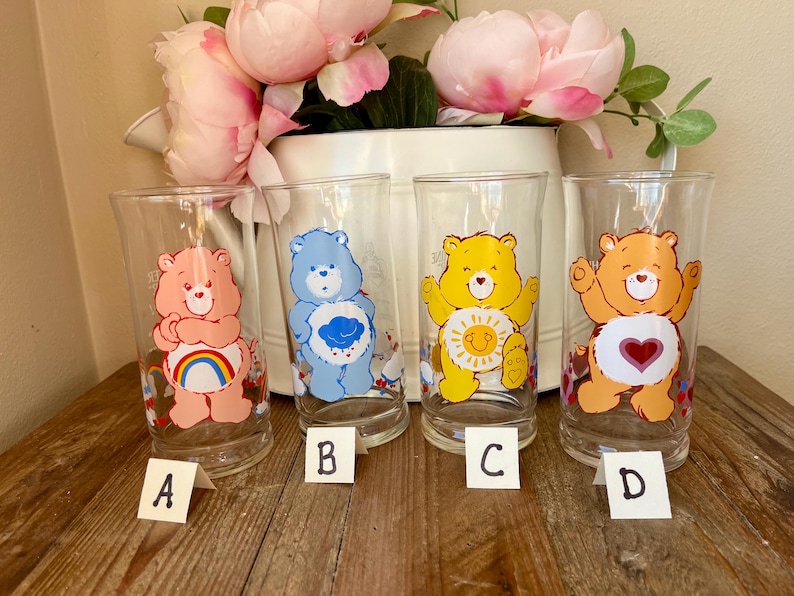 Op de afbeelding: Vier heldere glazen bekers met Care Bear-ontwerpen. Elk glas heeft een andere beer: roze met een regenboog, blauw met een wolk, geel met een zon en oranje met een hart. De glazen zijn gelabeld A, B, C en D.