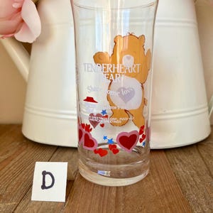 Op de afbeelding: Transparant glas met een cartoon berenontwerp, met de tekst "Tenderheart Bear" en "Share some love". Het glas is versierd met rode harten en blauwe sterren. Een witte kaart met de letter "D" staat voor het glas.