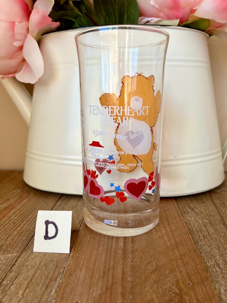 Op de afbeelding: Transparant glas met een cartoon berenontwerp, met de tekst "Tenderheart Bear" en "Share some love". Het glas is versierd met rode harten en blauwe sterren. Een witte kaart met de letter "D" staat voor het glas.