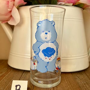 Op de afbeelding: Een helder glas met een lichtblauwe Care Bear-figuur met een wolkbuik en regenboogaccenten. Het glas is cilindrisch en staat op een houten oppervlak. Een witte kan met roze bloemen staat op de achtergrond.