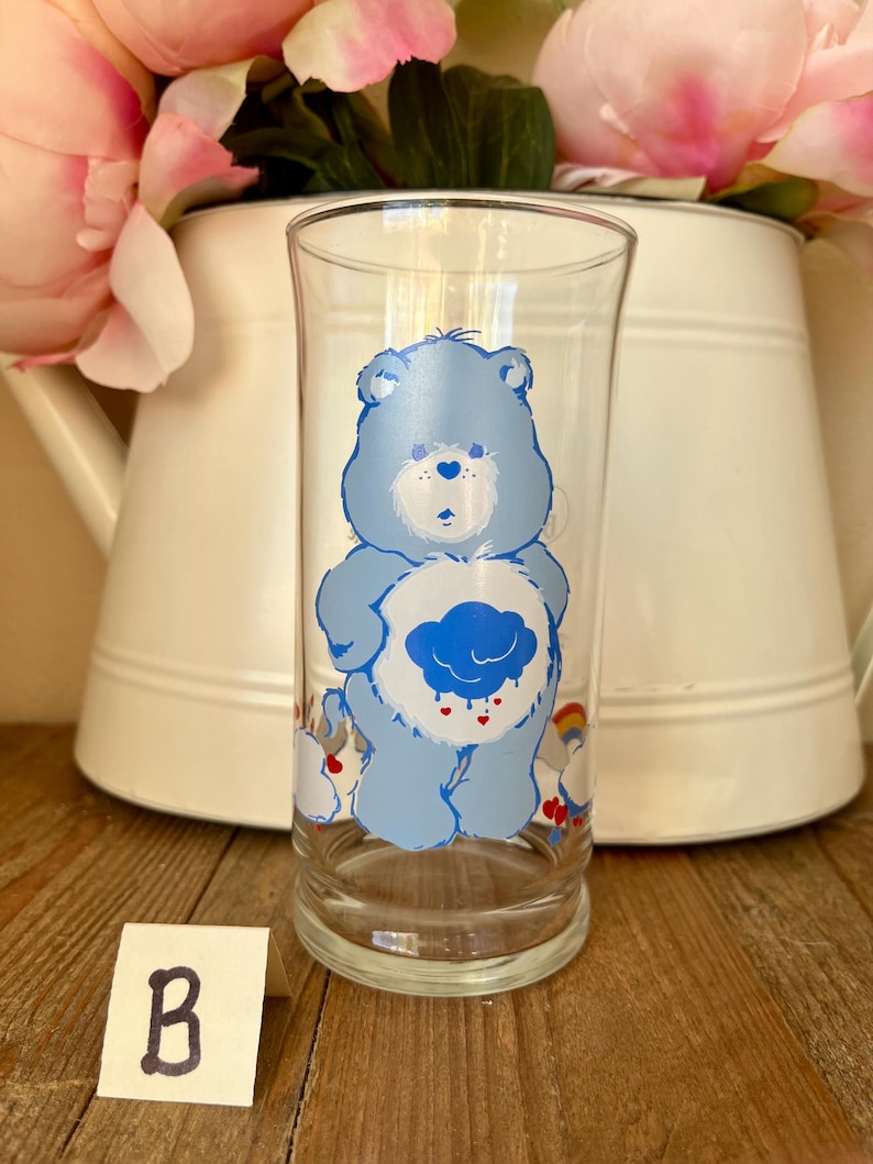 Op de afbeelding: Een helder glas met een lichtblauwe Care Bear-figuur met een wolkbuik en regenboogaccenten. Het glas is cilindrisch en staat op een houten oppervlak. Een witte kan met roze bloemen staat op de achtergrond.