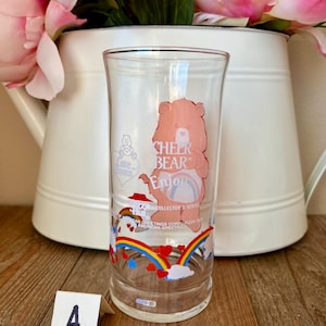Op de afbeelding: Een helder glas met een Cheer Bear cartoonontwerp en de tekst "CHEER BEAR Enjoy". Het glas heeft een regenboog- en hartontwerp aan de onderkant. Onderdeel van de Collector's Series van American Greetings Corp.