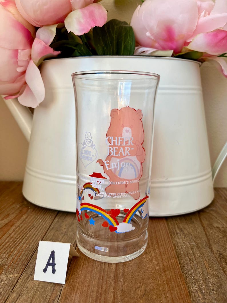 Op de afbeelding: Een helder glas met een Cheer Bear cartoonontwerp en de tekst "CHEER BEAR Enjoy". Het glas heeft een regenboog- en hartontwerp aan de onderkant. Onderdeel van de Collector's Series van American Greetings Corp.