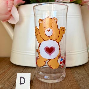 Op de afbeelding: Transparante glazen beker met een oranje berenontwerp en een rood hart op de buik. De beer lacht met zijn armen omhoog. Het glas staat tegen een witte achtergrond met roze bloemen en een witte kan.
