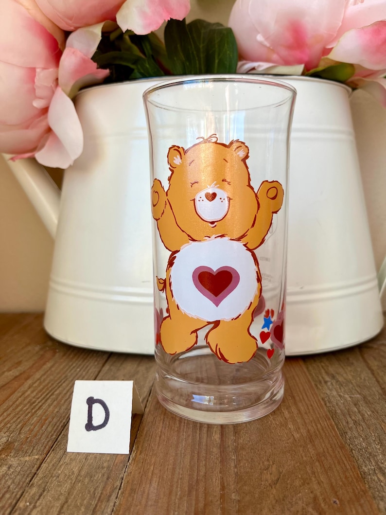 Op de afbeelding: Transparante glazen beker met een oranje berenontwerp en een rood hart op de buik. De beer lacht met zijn armen omhoog. Het glas staat tegen een witte achtergrond met roze bloemen en een witte kan.