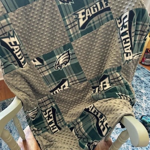 May include: A green and gray fleece blanket with a patchwork design featuring the Philadelphia Eagles logo and the words "Eagles" repeated throughout. The blanket is folded and draped over a wooden chair.